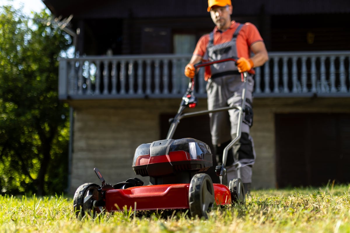 A man in work clothes pushes a cordless lawn mower across a residential lawn during home garden maintenance
