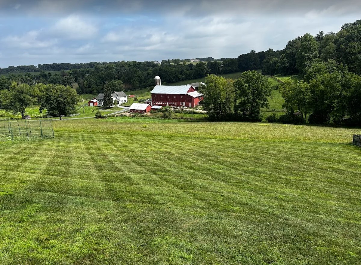 Rural farmland with red barn and white house surrounded by green fields and forested hills under partly cloudy sky