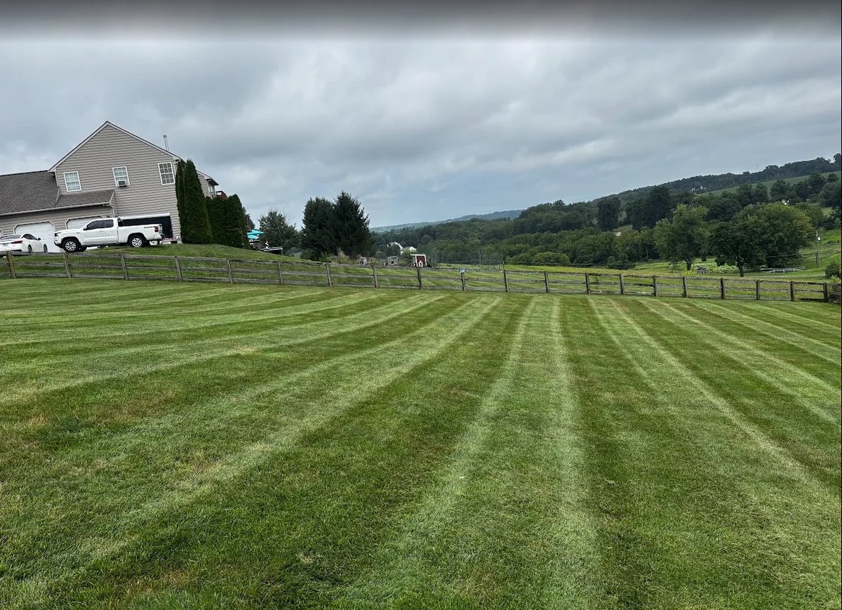 A freshly mowed farm field with striped grass patterns, gray barn with vehicles, and forested hills under cloudy skies